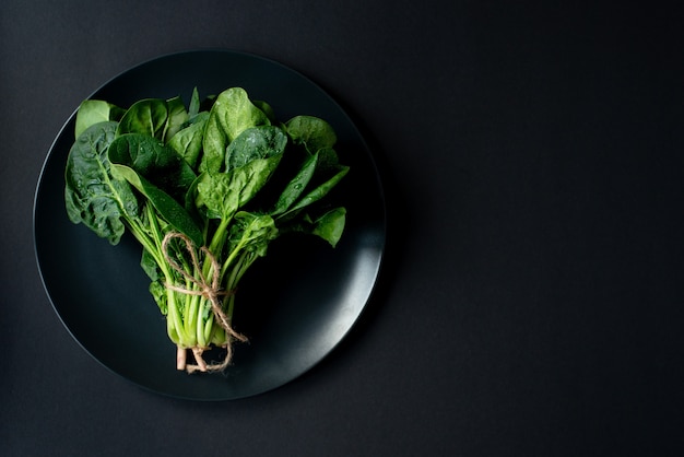 Schoon voedselconcept. Bos van bladeren van verse biologische spinazie greens in een plaat op een zwarte achtergrond. Gezond detox lente-zomer dieet. Veganistisch rauw voedsel. Kopieer ruimte.