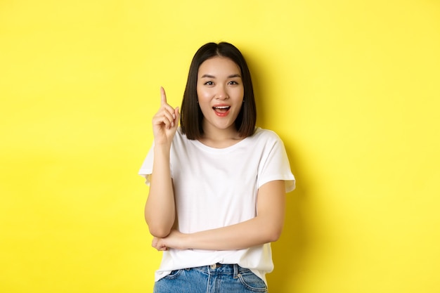 Schoonheid en mode concept. Opgewonden Aziatisch meisje dat vinger opsteekt in eureka-gebaar, een idee gooit en glimlacht, staande over gele achtergrond