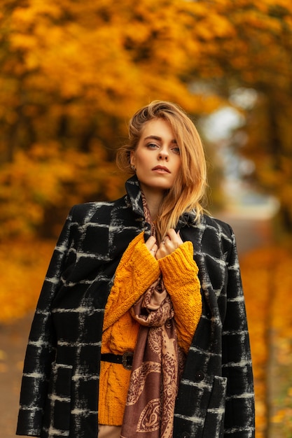 Schoonheid Europees modelmeisje in mode-herfstkleding met zwarte jas, vintage trui en sjaal loopt in een geweldig geel park met herfstbladeren