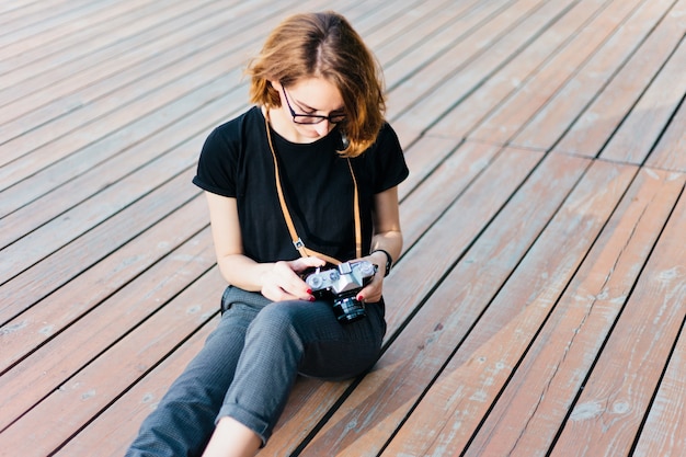 Schoonheid hipster vrouw met bril maakt gebruik van retro camera zittend op houten vloer buitenshuis