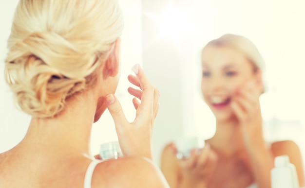 schoonheid, huidverzorging en mensenconcept - close-up van een glimlachende jonge vrouw die crème aanbrengt op het gezicht en kijkt naar een spiegel in de badkamer thuis