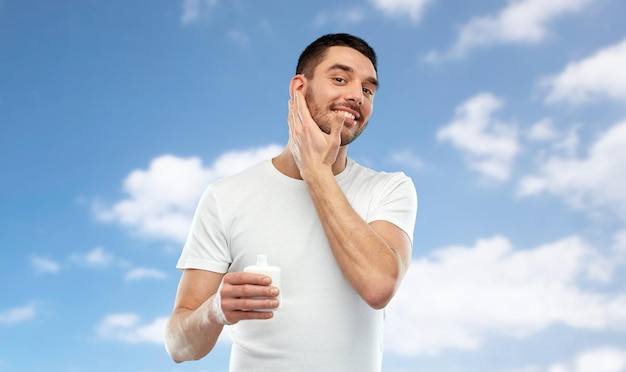 schoonheid, huidverzorging, lichaamsverzorging en mensenconcept - lachende jonge man die crème of lotion aanbrengt op de blauwe lucht en de wolken achtergrond