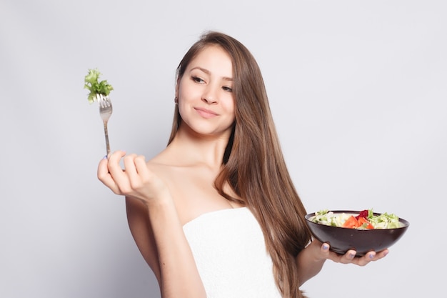 Schoonheid, huidzorg en mensenconcept - mooi donkerbruin meisje die in handdoek verse salade eten en glimlachen. vrouw die voedsel eet. mooie glimlachende vrouw met voor het diner verse groenten in de keuken.