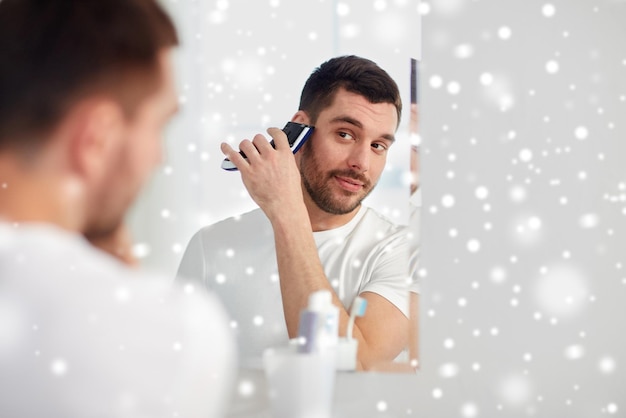 schoonheid, hygiëne, scheren, verzorging en mensenconcept - jonge man op zoek naar spiegel en scheerbaard met trimmer of elektrisch scheerapparaat thuis badkamer over sneeuw