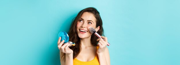 Schoonheid jong glamourmeisje dat make-up aanbrengt met zakspiegel en borstel glimlachend en starend naar log