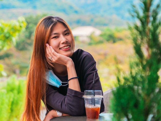 Foto schoonheid jonge aziatische lachende vrouw zit in cafe met bos natuur achtergrond met ijskoffie op tafel