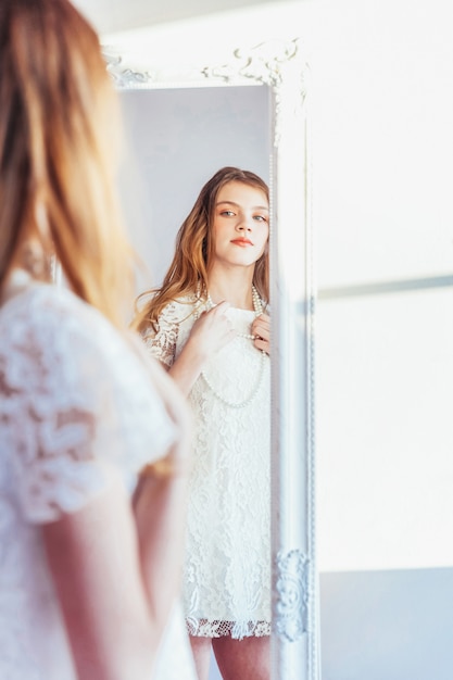 Schoonheid make-up ochtend rutine hou van jezelf concept. jonge tiener die bezinning in spiegel bekijkt. jonge positieve vrouw die het witte kleding stellen in heldere lichte ruimte dragen tegen witte muur.