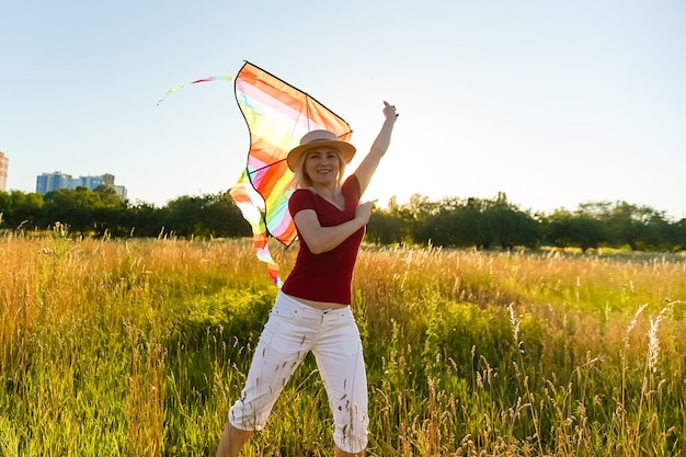 Schoonheid meisje loopt met vlieger op het veld. Mooie jonge vrouw met vliegende kleurrijke vlieger over heldere blauwe hemel. Gratis, vrijheidsconcept. Emoties, gezonde levensstijl