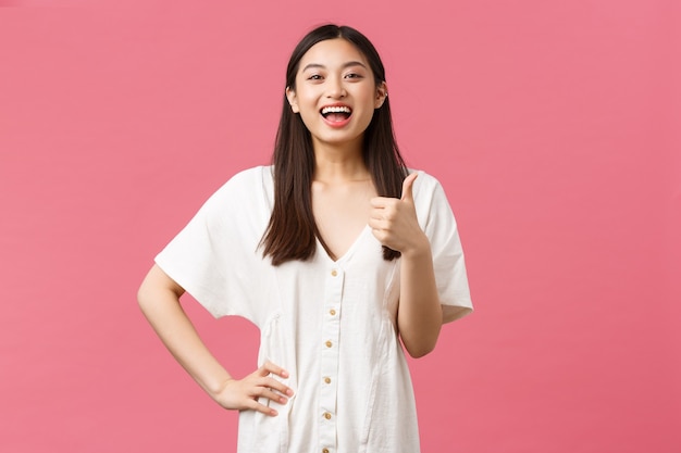 Schoonheid, mensen emoties en zomer vrije tijd en vakantie concept. Blije, tevreden Aziatische vrouwelijke klant in de winkel, duim omhoog en vrolijk lachend, bezoekplaats aanbevelen, roze achtergrond