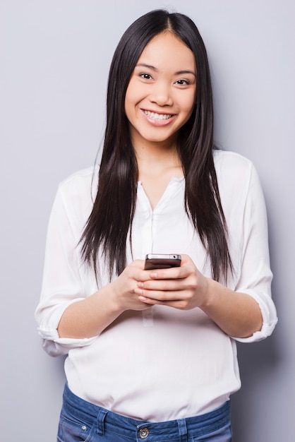 Schoonheid met telefoon. Vrolijke jonge Aziatische vrouw die mobiele telefoon vasthoudt en glimlacht terwijl ze tegen een grijze achtergrond staat