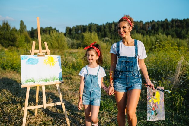 Schoonheid moeder schildert met haar dochtertje. Stijlvolle vrouw die de afbeelding met een klein meisje tekent. Schattige jongen in een wit t-shirt en spijkerbroek.