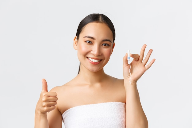 Schoonheid, persoonlijke en intieme zorg menstruele hygiëne concept. Close-up van mooie jonge Aziatische vrouw in badhanddoek die tampon en thumbs-up tonen, die op haar peirod, witte achtergrond zijn.