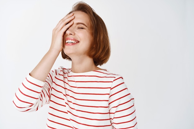 Schoonheid. Portret van een gelukkige mooie dame die zorgeloos lacht en glimlacht, de helft van het gezicht bedekt met de hand, met gesloten ogen tegen een witte muur staat