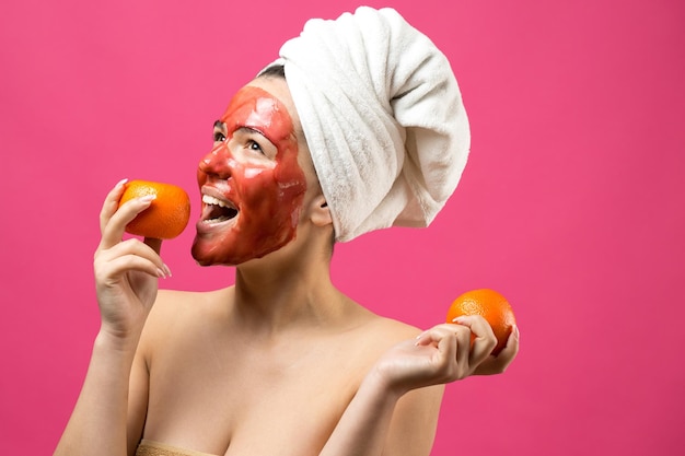 Schoonheid portret van vrouw in witte handdoek op hoofd met rood voedend masker op gezicht Huidverzorging reiniging eco organische cosmetische spa ontspannen concept Een meisje staat met haar rug met een oranje mandarijn