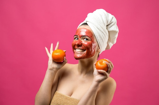 Schoonheid portret van vrouw in witte handdoek op hoofd met rood voedend masker op gezicht Huidverzorging reiniging eco organische cosmetische spa ontspannen concept Een meisje staat met haar rug met een oranje mandarijn