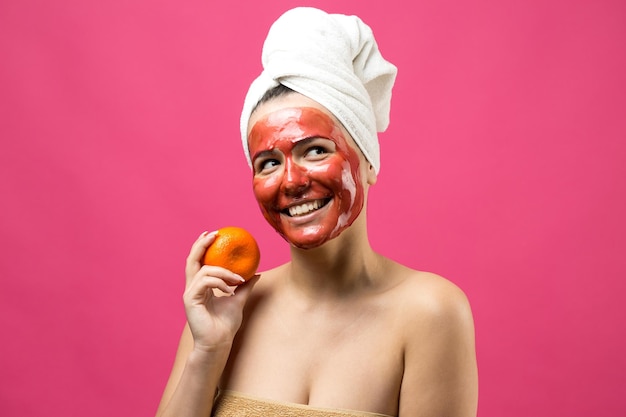 Schoonheid portret van vrouw in witte handdoek op hoofd met rood voedend masker op gezicht Huidverzorging reiniging eco organische cosmetische spa ontspannen concept Een meisje staat met haar rug met een oranje mandarijn