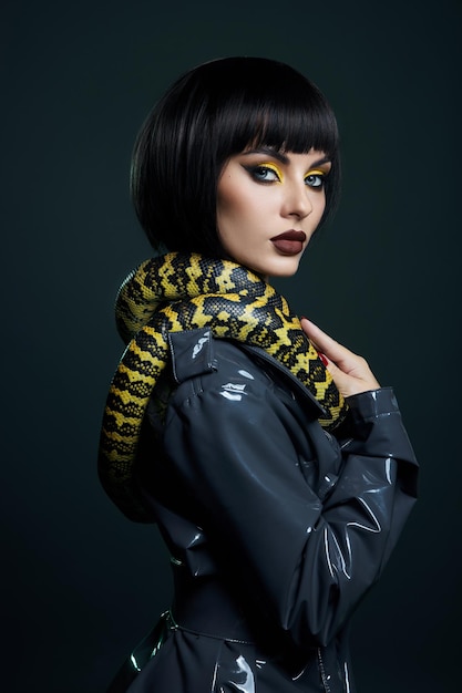 Schoonheid vrouw python gele slang rond haar nek op latex glanzende regenjas. Gele slang op de schouders van meisje. Schoonheid gele oogschaduw make-up, donkere bordeauxrode lippenstift