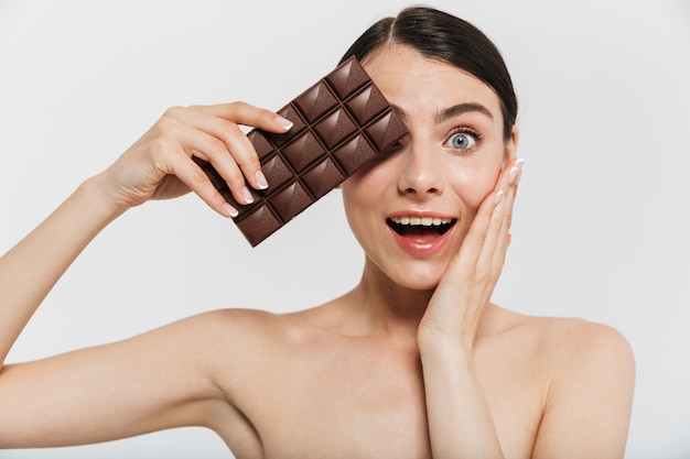 Schoonheidsportret van een aantrekkelijke jonge donkerbruine vrouw die zich geïsoleerd over witte muur bevindt, die zwarte chocoladereep toont