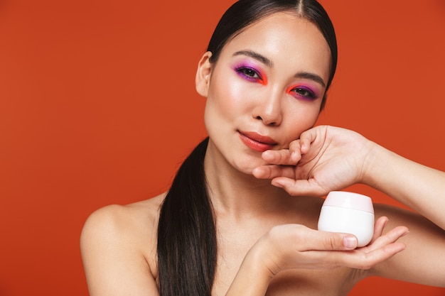 Schoonheidsportret van een gelukkige jonge topless aziatische vrouw met donkerbruin haar die lichte make-up draagt, geïsoleerd op rood staat, fles met gezichtscrème toont