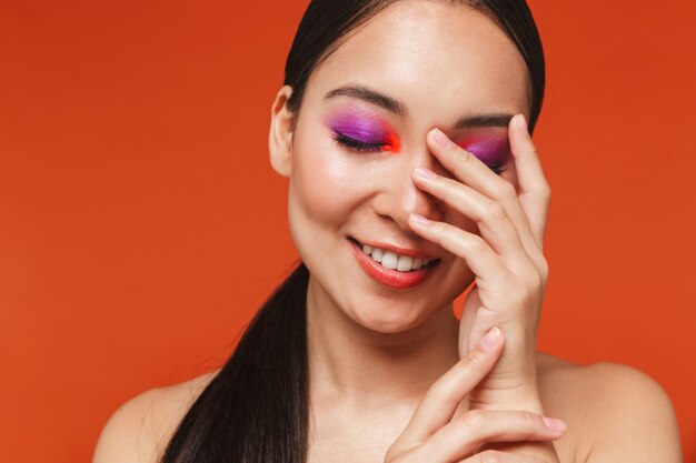 Schoonheidsportret van een gelukkige jonge topless aziatische vrouw met donkerbruin haar die lichte make-up draagt, geïsoleerd op rood staat