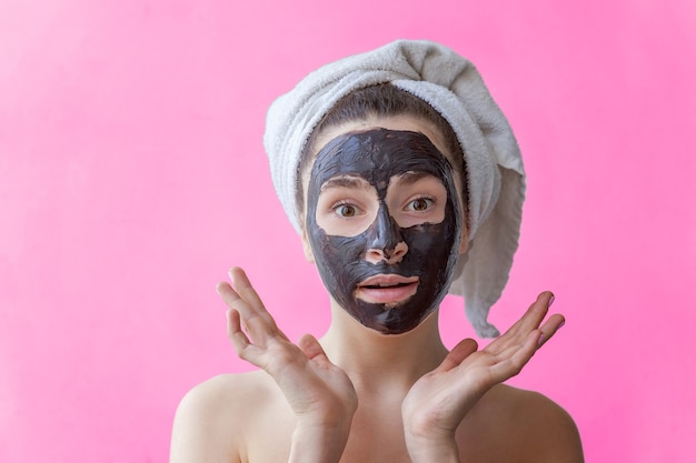 Schoonheidsportret van een glimlachende donkerbruine vrouw in een handdoek op het hoofd die zwart voedend masker op gezicht op roze geïsoleerde achtergrond toepast. Huidverzorging reiniging spa ontspannen cosmetica concept