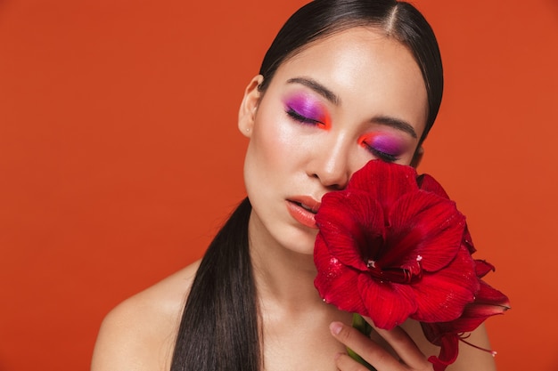 Schoonheidsportret van een jonge topless aziatische vrouw met donkerbruin haar die lichte make-up draagt, geïsoleerd op rood staat, poserend met een rode bloem