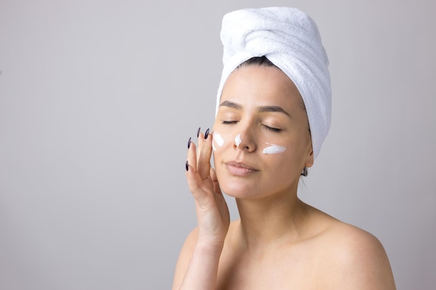 Schoonheidsportret van een vrouw in een witte handdoek op het hoofd brengt crème aan op het gezicht Huidverzorging reiniging eco-organische cosmetische spa relax concept