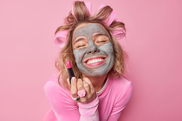 Schoonheidsportret van gelukkige Europese vrouw past schoonheidsmasker toe op gezicht houdt lippenstift gebruikt cosmetisch product past haarrollers toe en bereidt zich voor op datum geïsoleerd over roze achtergrond Make-uproutine