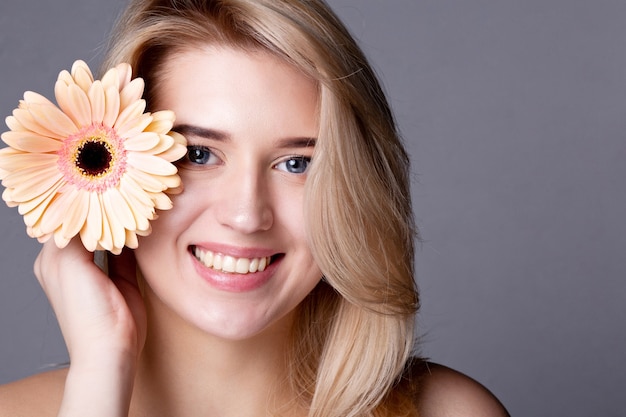 Schoonheidsportret van luxe blond model met naakte schouders die zich voordeed in studio met roze gerbera. Ruimte voor tekst