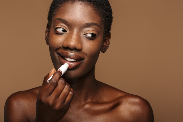 Schoonheidsportret van mooie jonge halfnaakte Afrikaanse vrouw die lippenbalsem opdoet die op bruin wordt geïsoleerd