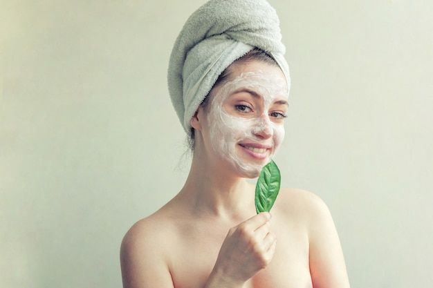 Schoonheidsportret van vrouw in handdoek op hoofd met wit voedend masker of room op gezicht en groen blad ter beschikking, witte geïsoleerde achtergrond. Huidverzorgingsreiniging eco biologisch cosmetisch spa-concept