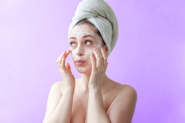 Schoonheidsportret van vrouw in handdoek op hoofd met wit voedend masker of room op gezicht, purpere geïsoleerde achtergrond