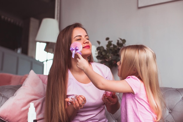 Schoonheidssalon thuis Meisje doet make-up op haar moeders gezicht met roze zachte spons Moeder en dochter spelen schoonheidssalon tijdens vakantie of vakantie