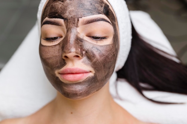 Schoonheidsspecialist die een zwart masker toepast op het gezicht van een mooie vrouw met zwarte handschoenen, een prachtige vrouw in de spa met gezichtsprocedures