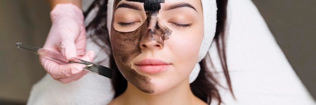 Schoonheidsspecialist die een zwart masker toepast op het gezicht van een mooie vrouw met zwarte handschoenen, een prachtige vrouw in de spa met gezichtsprocedures