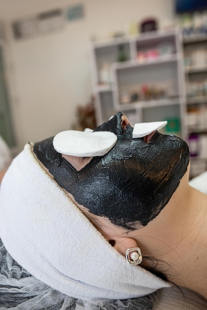 Schoonheidsspecialiste brengt een zwart masker aan op het gezicht van haar cliënt in een schoonheidssalon Concept van huidreiniging en hydratatie