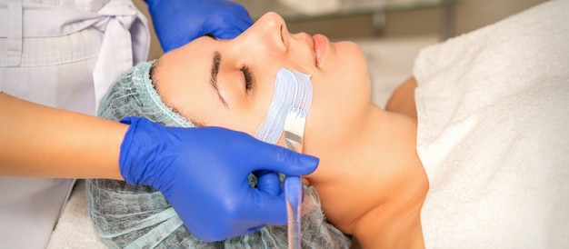 Schoonheidsspecialiste die de gezichtshuid van de vrouw bedekt met een hydraterend reinigingsmasker tijdens de huidverzorgingsprocedure