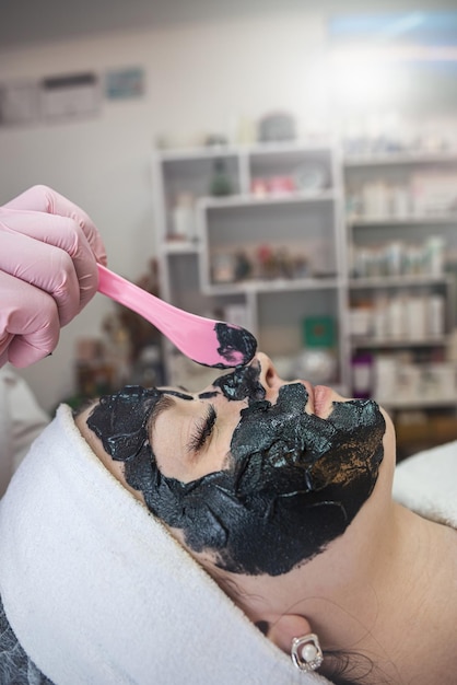 Schoonheidsspecialiste die zwart masker toepast op het gezicht van een jonge mooie vrouw in de spa salon