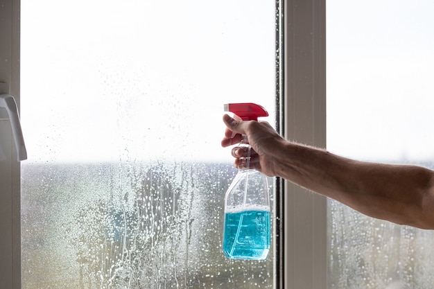 Schoonmaakdienst kwam om nieuw huis schoon te maken. Hardwerkende blanke man reinigt zorgvuldig raam. Man rode handschoenen houden spuiten blauwe fles op raam. Ramen reinigen met speciale doek en reiniger