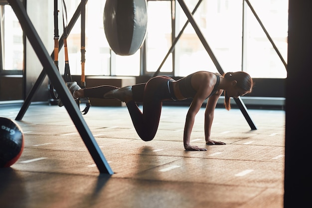 Schorsing opleiding. Zijaanzicht van jonge blanke fitnessvrouw in sportkleding die TRX-training doet in de industriële sportschool, trainen met fitnessriemen. Sport, wellness en een gezonde levensstijl