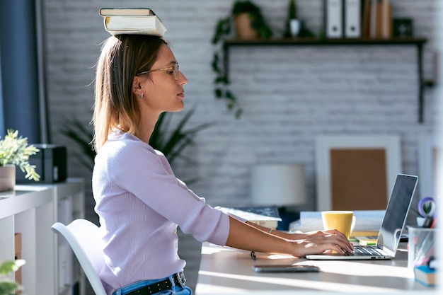 Schot van jonge aantrekkelijke vrouw aan de balie met boeken op haar hoofd tijdens het werken met de computer thuis.