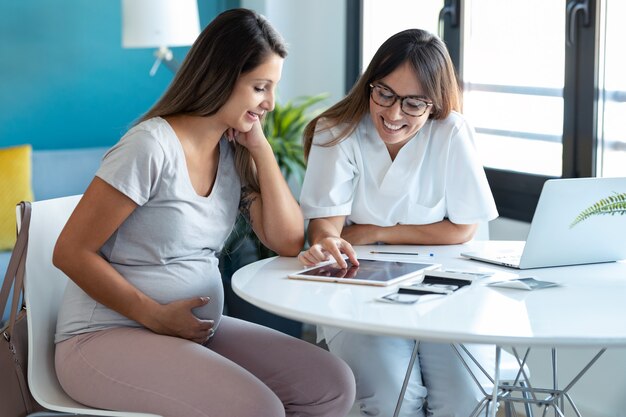 Schot van jonge vrouw gynaecoloog arts die in medisch overleg aan zwangere vrouw echografie baby met digitale tablet laat zien.