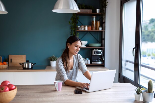 Schot van lachende jonge vrouw die met laptop in de keuken thuis werkt.