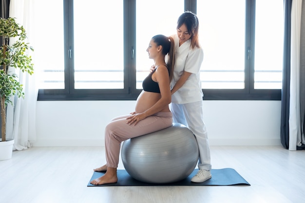 Schot van mooie jonge fysiotherapeut die mooie zwangere vrouw helpt voor het doen van pilates-oefeningen met bal die zich thuis voorbereidt op de bevalling.