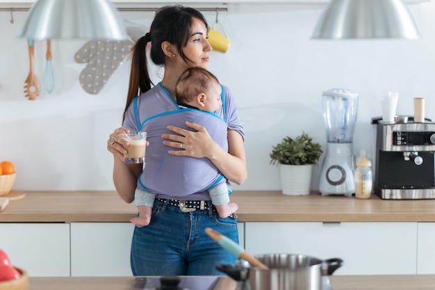 Schot van mooie jonge moeder met kleine baby in draagdoek koffie drinken in de keuken thuis.