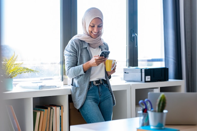 Schot van mooie jonge moslim zakenvrouw die hijab draagt met haar smartphone terwijl ze naast het raam op kantoor staat.