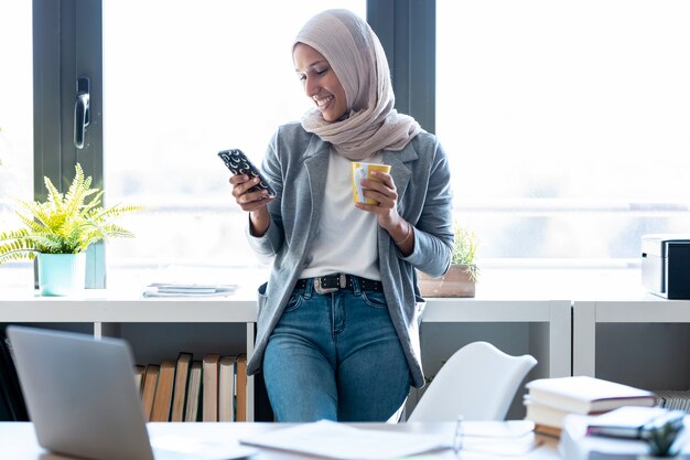 Schot van mooie jonge moslim zakenvrouw die hijab draagt met haar smartphone terwijl ze naast het raam op kantoor staat.