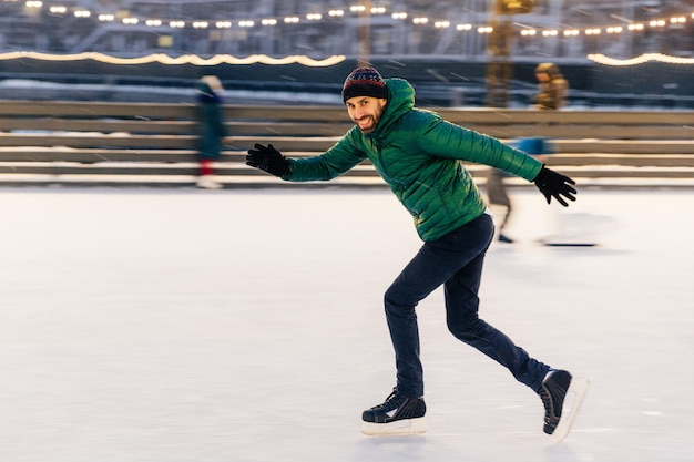 Schot van vrolijke man heeft plezier buiten schaatsen op ijsring geniet van deze winteractiviteit beweegt actief probeert evenwicht te bewaren goed getraind is kijkt naar de camera met gelukkige uitdrukking