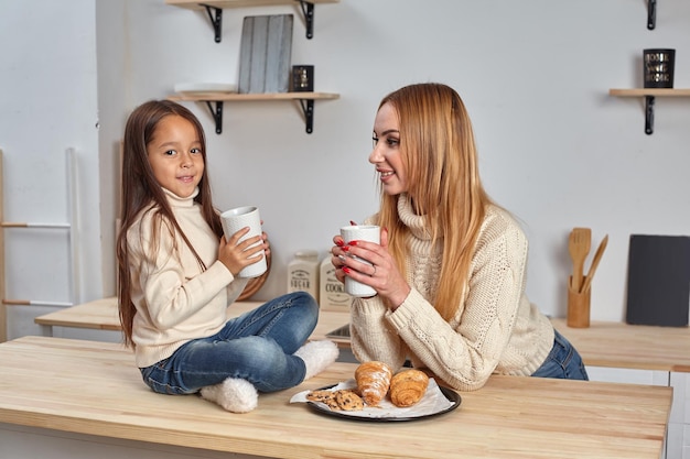 Schot van vrolijke moeder en dochter zitten samen aan de keukentafel en drinken 's ochtends hete thee.