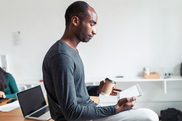 Schot van zwarte jonge zakenman die met digitale tablet werkt terwijl hij koffie drinkt op kantoor.